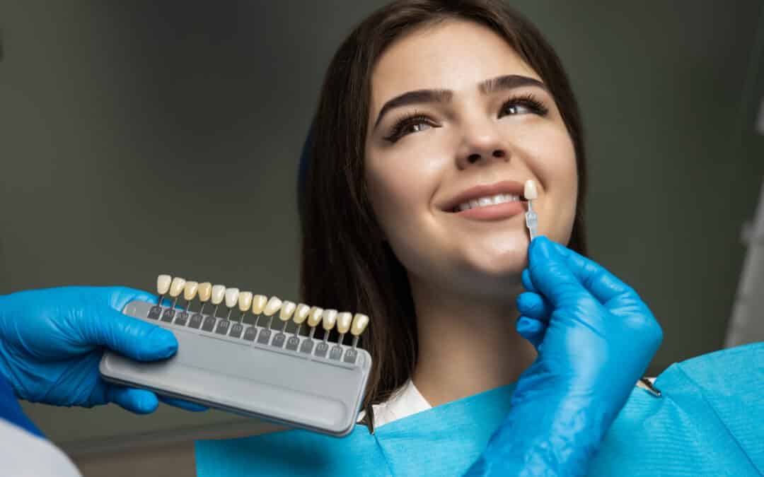 dentist in blue medical gloves applying sample from tooth enamel scale to happy woman patient teeth to pick up right shade for teeth veneer procedure